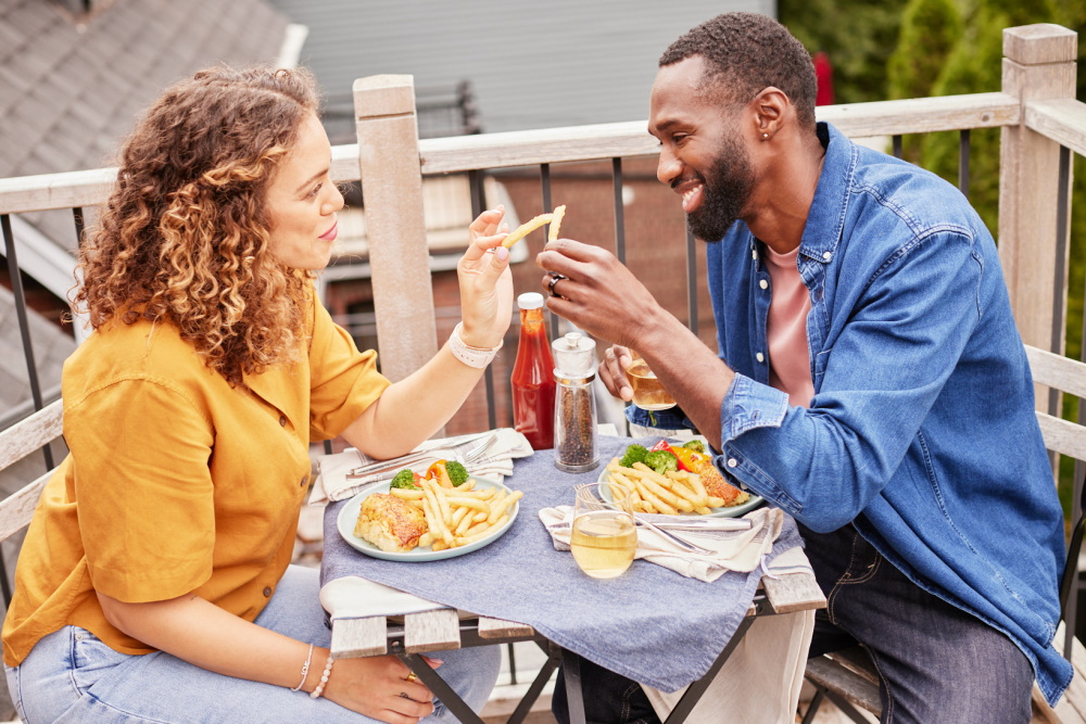 Couple sitting at a bistro table for two on their patio toasting their meal with their fries. 